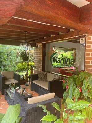 A covered outdoor seating area with wicker furniture, a glass wall with the La Martina Hotel Campestre logo, and lush greenery in the background. - Hotel Campestre La Martina (Rionegro)