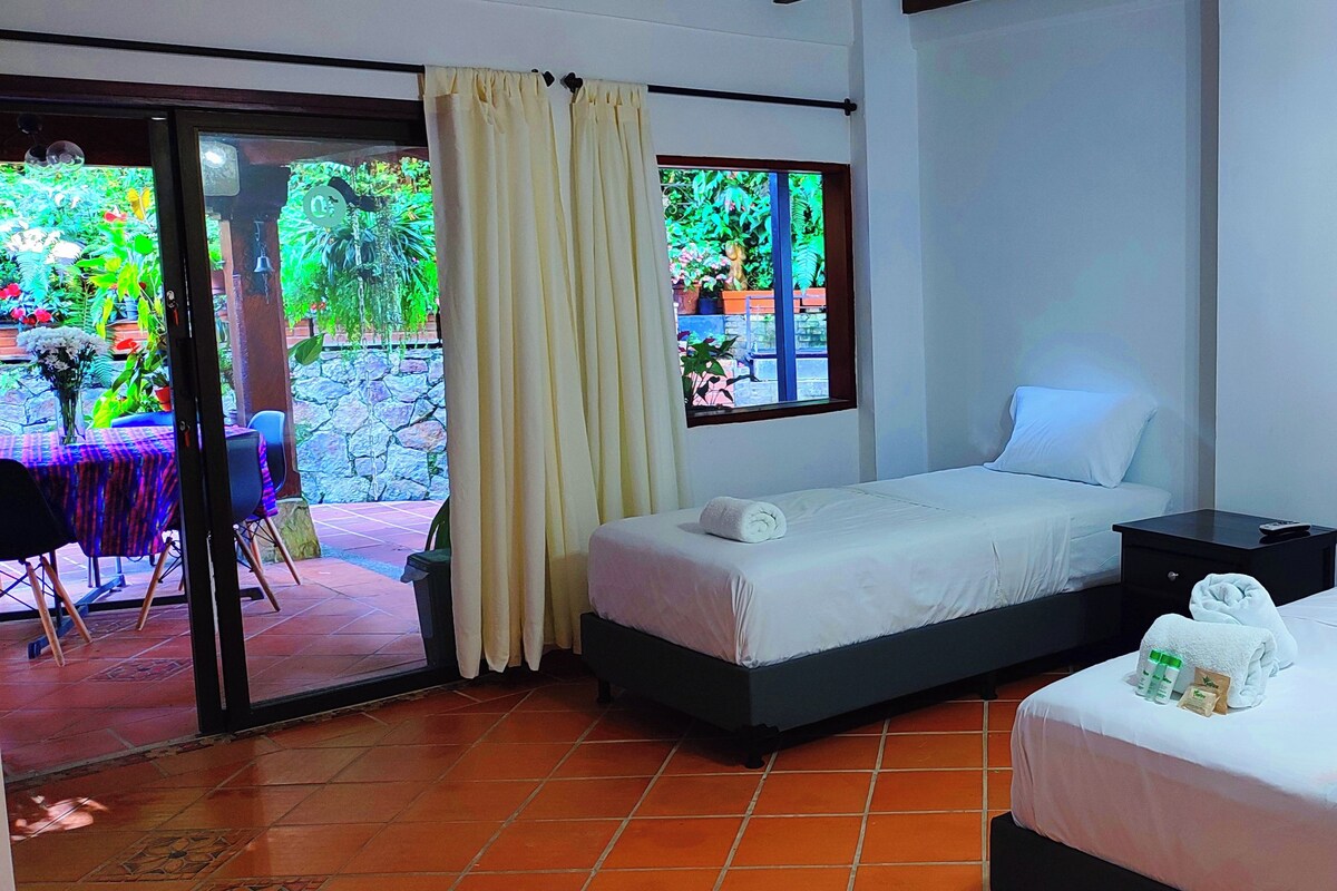 A hotel room with two beds, a small table, and a view of a garden through a window.