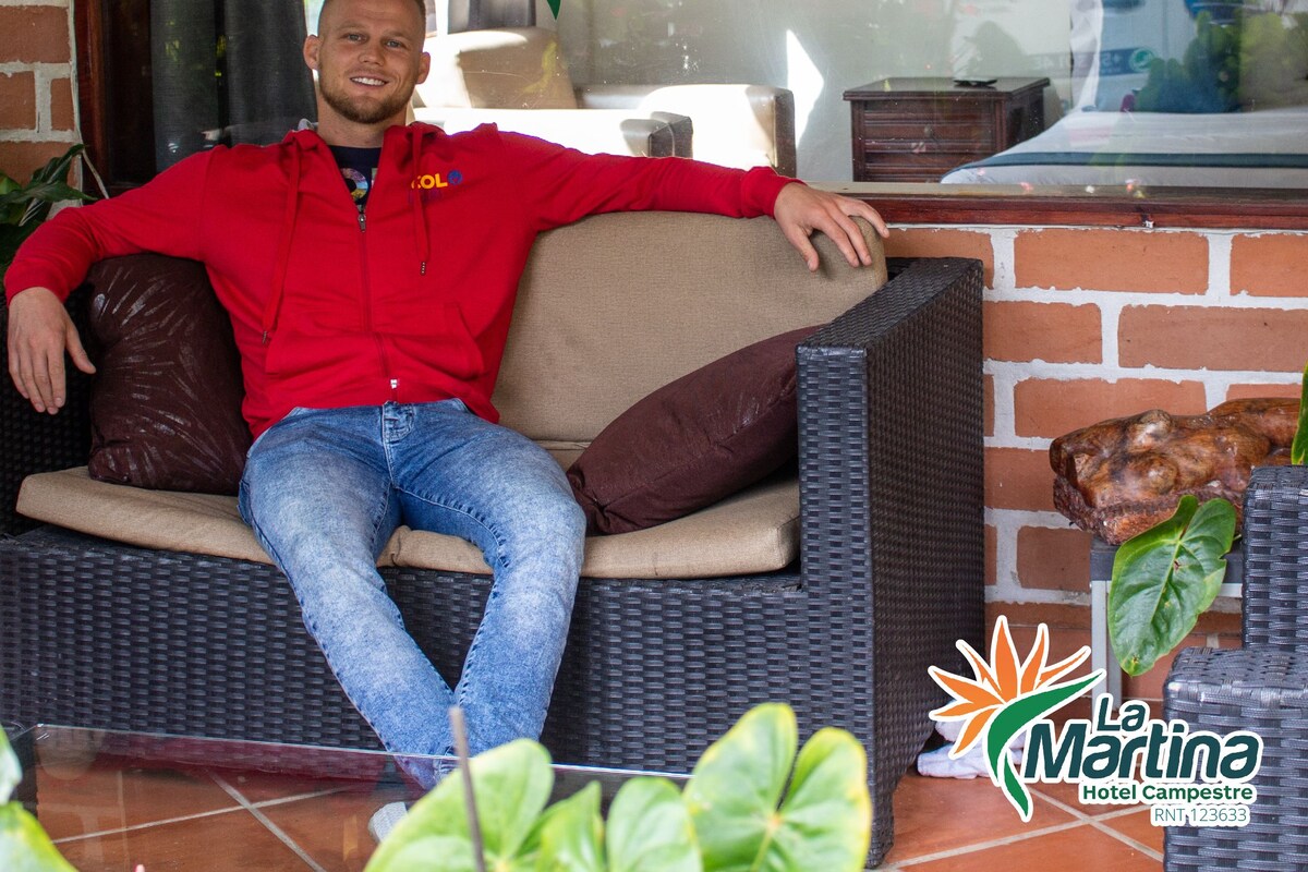 A man sitting on a wicker sofa with a cushion, in front of a sign for La Martina Hotel Campestre.