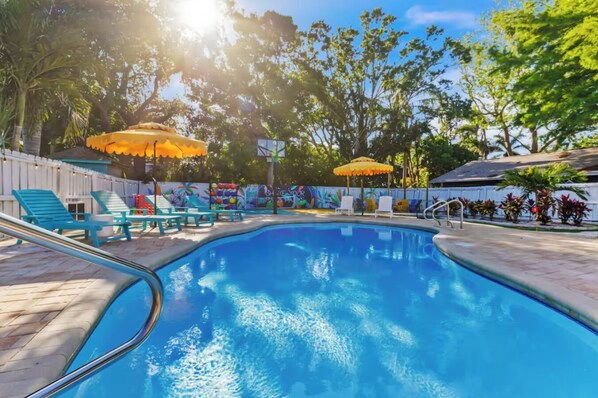 A swimming pool with lounge chairs and umbrellas, surrounded by trees and a white fence.