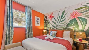 Ein Hotelzimmer mit Bett, Nachttisch, Lampe und Wandbild mit tropischem Motiv.