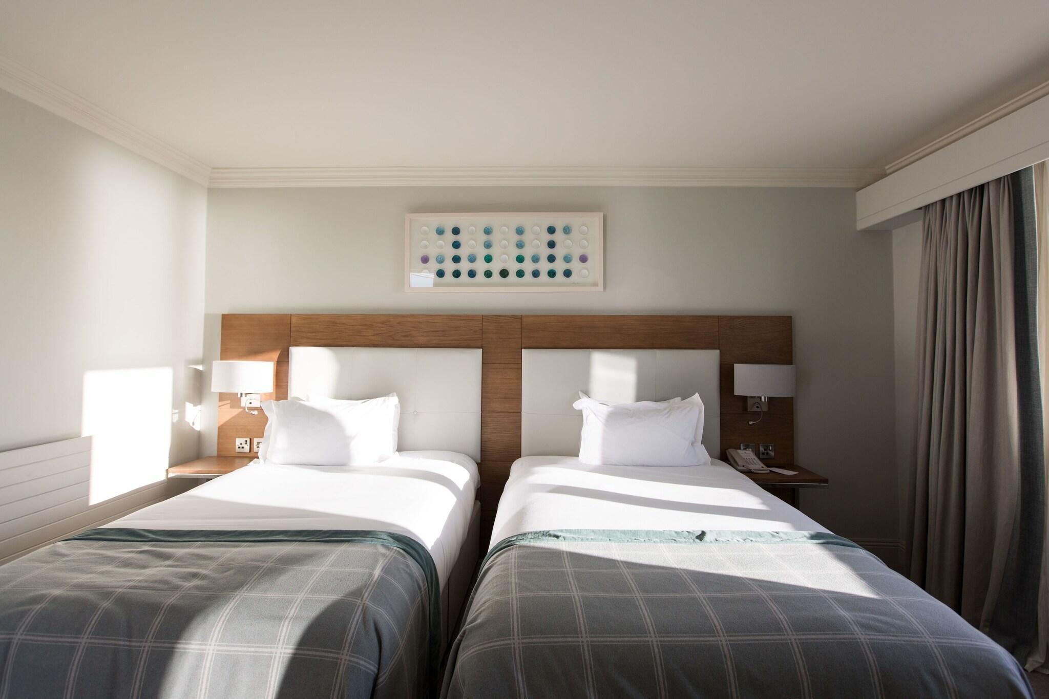 Habitación de hotel con dos camas, cabeceros, mesitas de noche con lámparas, un teléfono y una obra de arte en la pared.