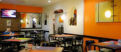 A restaurant with multiple tables and chairs, a television mounted on the wall, and a ceiling with a floral design.