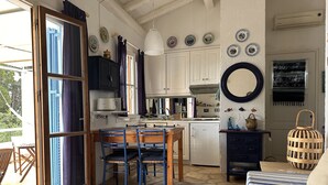 Une cuisine avec des meubles blancs, une table à manger avec des chaises bleues, un réfrigérateur et un sol en pierre.