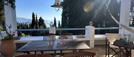 An outdoor patio with a wooden table and chairs, a hanging light fixture, and a view of the sea and mountains.