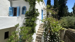 Un bâtiment blanc avec des volets bleus et un escalier en pierre menant à une porte. De grands cyprès sont visibles à l’arrière-plan.