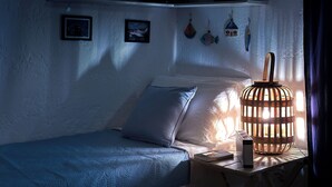 Une chambre à coucher faiblement éclairée, avec un lit simple, une table de chevet avec une lampe, et des éléments de décoration murale, notamment des tableaux encadrés et des ornements suspendus.