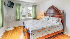 Un dormitorio con cama de madera, cortinas verdes, ventilador de techo, televisor montado en la pared y ventana con vistas al exterior.