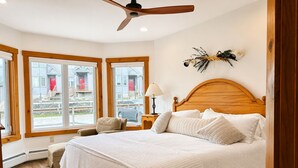 Een slaapkamer met een houten hoofdeinde, een bed met witte lakens, een stoel, een nachtkastje met een lamp en een raam met uitzicht op gebouwen.