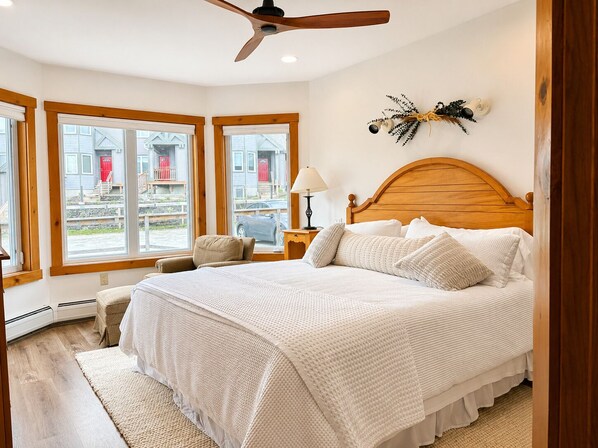 Un dormitorio con cabecera de madera, cama con ropa de cama blanca, una silla, una mesita de noche con lámpara y ventana con vistas a edificios.