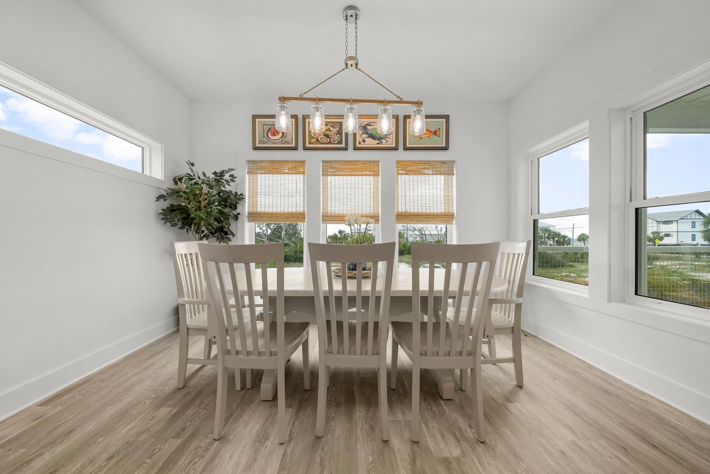 Un coin repas avec une table et des chaises blanches, un luminaire suspendu et de grandes fenêtres équipées de stores.