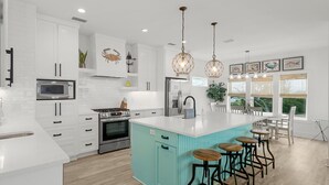 A modern kitchen with white cabinetry, a teal island, stainless steel appliances, and pendant lighting. - Beach House - Even Keel by PHG (Mexico Beach)