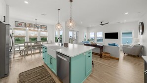 A modern kitchen with a central island, dining area with chairs, and a living room with a sofa and TV. - Beach House - Even Keel by PHG (Mexico Beach)