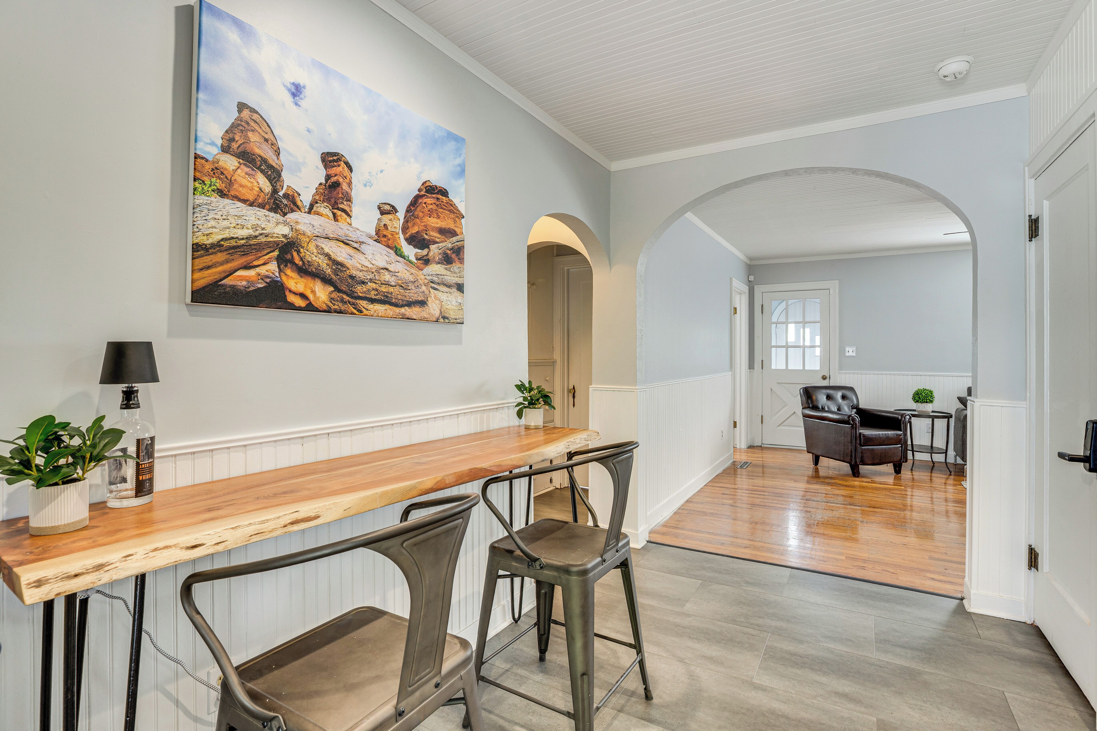 Un hall spacieux avec une table à manger en bois et deux tabourets, un tableau représentant des formations rocheuses accroché au mur, et une vue sur une autre pièce où l’on aperçoit une chaise et une table.