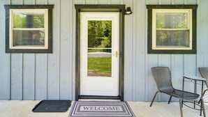 Une porte d’entrée avec un paillasson sur lequel est écrit « BIENVENUE », deux fenêtres avec des stores et une chaise à l’extérieur.