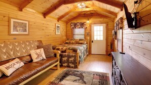 Ein gemütliches Blockhaus-Interieur mit Holzboden, einem Bett mit gemusterten Betttextilien, einem Ledersofa mit Hirsch-Kissen und einer Kommode.