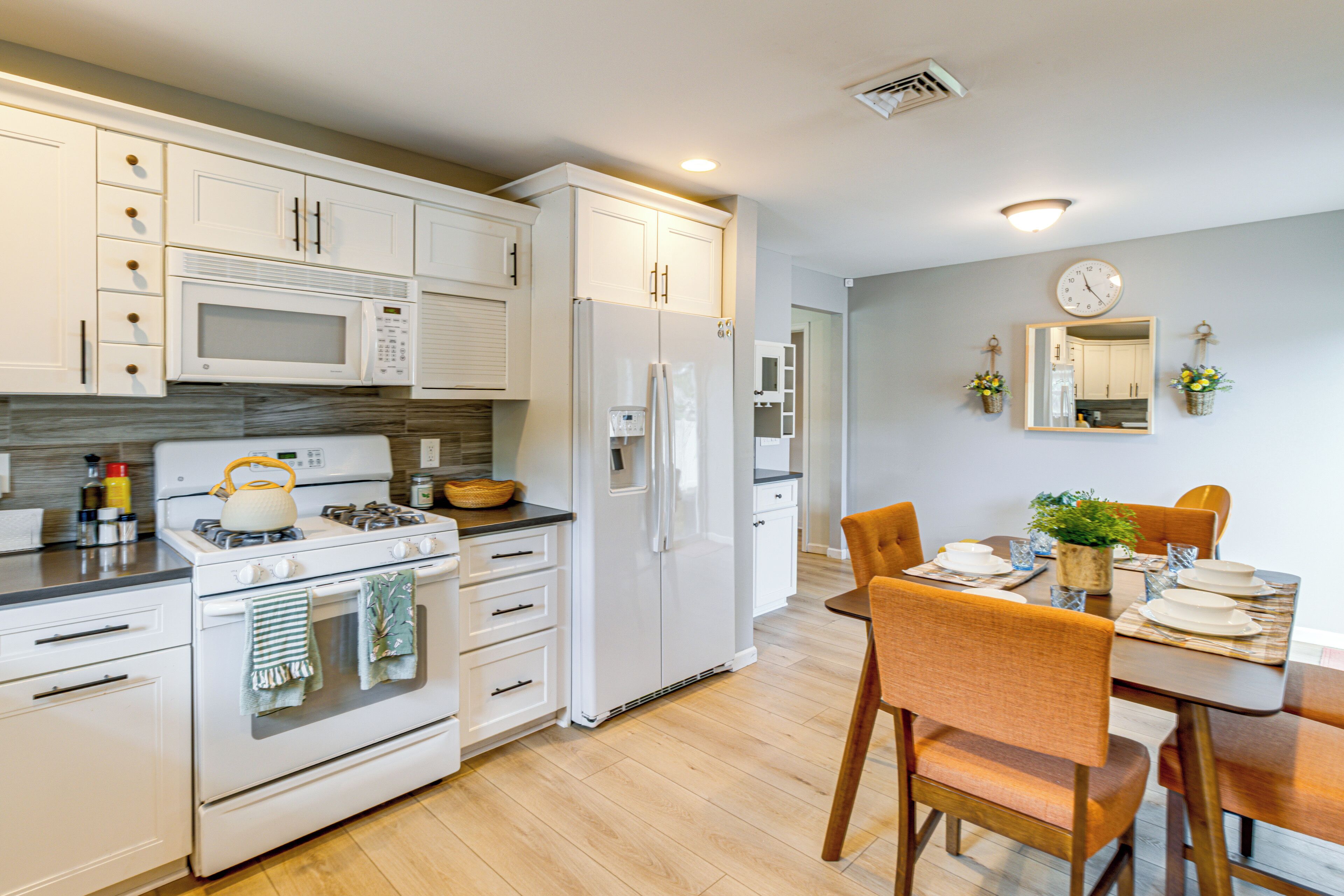 A modern kitchen with white cabinetry, a stove, microwave, and refrigerator. There is a dining table with chairs and a clock on the wall.