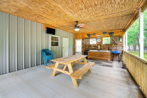 Ein Holztisch mit Bänken in einem überdachten Außenbereich, inklusive Deckenventilator, Küchenzeile und Blick ins Grüne.