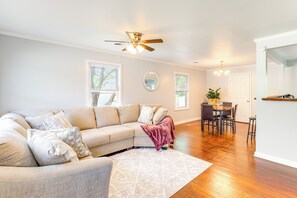 Un salon avec un canapé d’angle beige, un ventilateur de plafond et un coin repas comprenant une table et des chaises en bois.