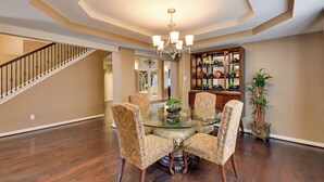 A dining area with a glass table surrounded by patterned chairs, a chandelier, and a wooden staircase in the background.