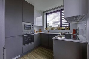 A modern kitchen with grey cabinets, a built-in oven, a microwave, and a window with a view of the city. - Arran Quay - Ardcairn House - Campus Accommodation (Dublin)