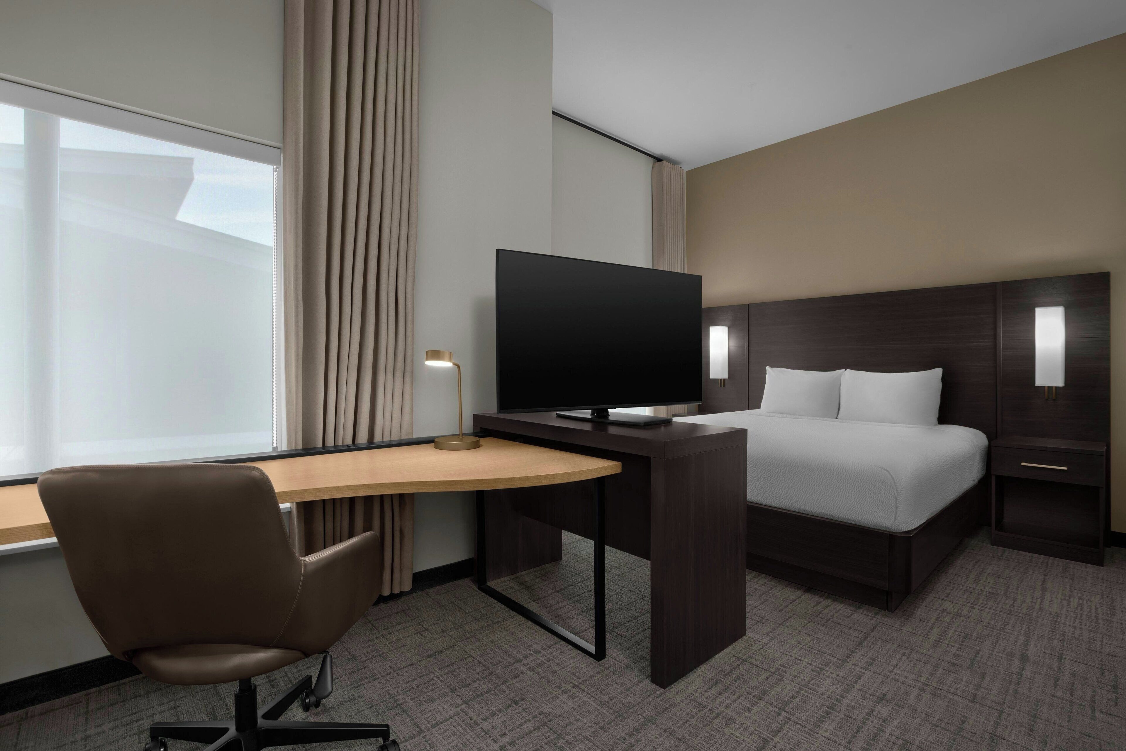 A hotel room with a bed, a desk with a TV, a chair, and a nightstand with a lamp.