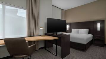 A hotel room with a bed, a desk with a TV, a chair, and a nightstand with a lamp.