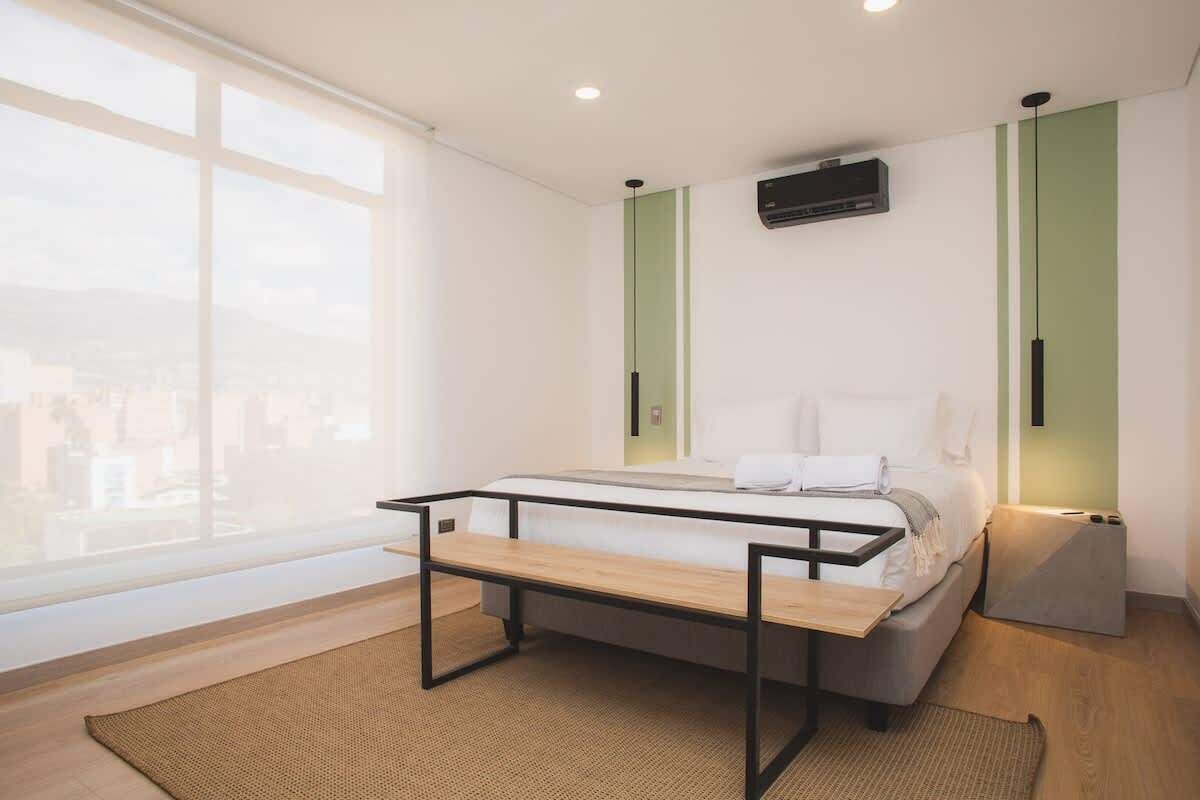 A modern bedroom with a bed, a small table, and a bench. There is a window with a view of buildings outside.