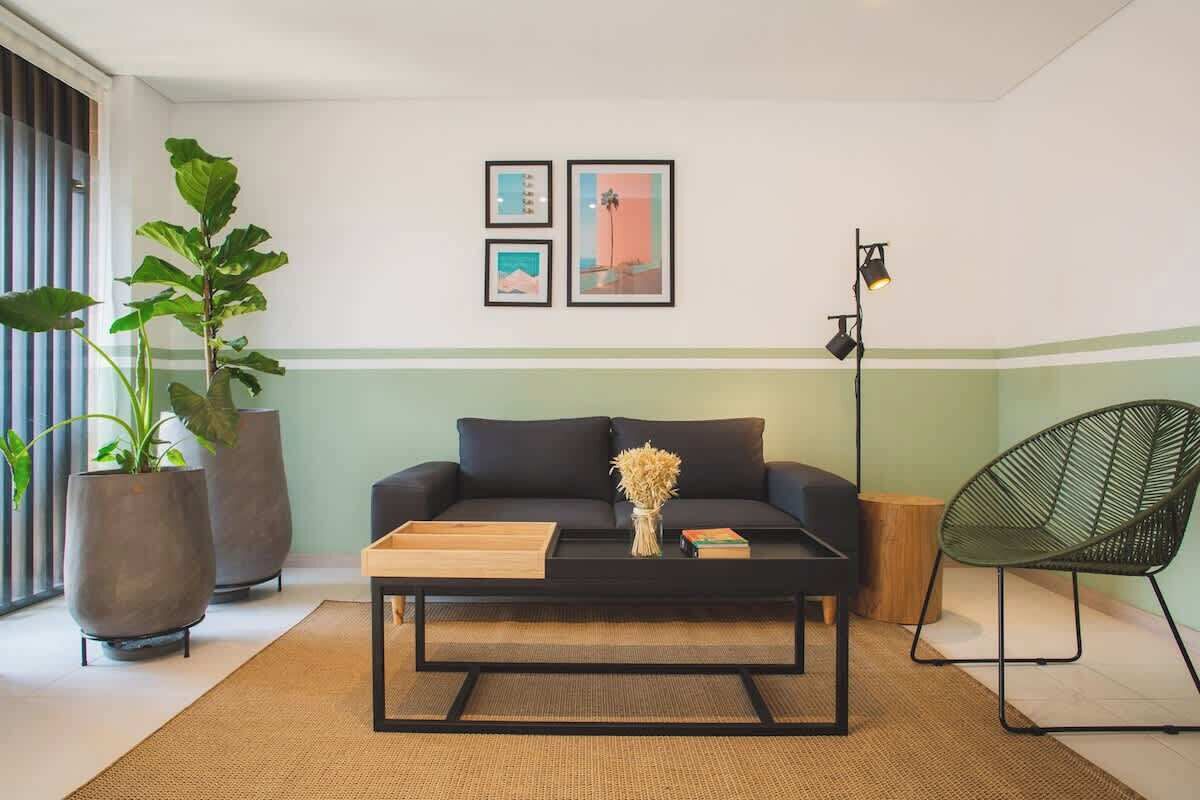 En moderne stue med en sort sofa, et sofabord og en grøn lænestol. To store potteplanter og tre billeder hænger på væggen.