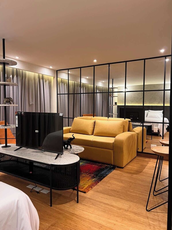 A modern hotel room with a yellow sofa, a flat-screen TV, a small round table, and a colorful rug.