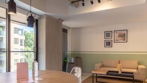 Ein modernes Wohnzimmer mit einem runden Holztisch, einer Couch und einem Couchtisch. Ein Fenster bietet Blick auf Gebäude, auf dem Tisch steht eine Pflanze.