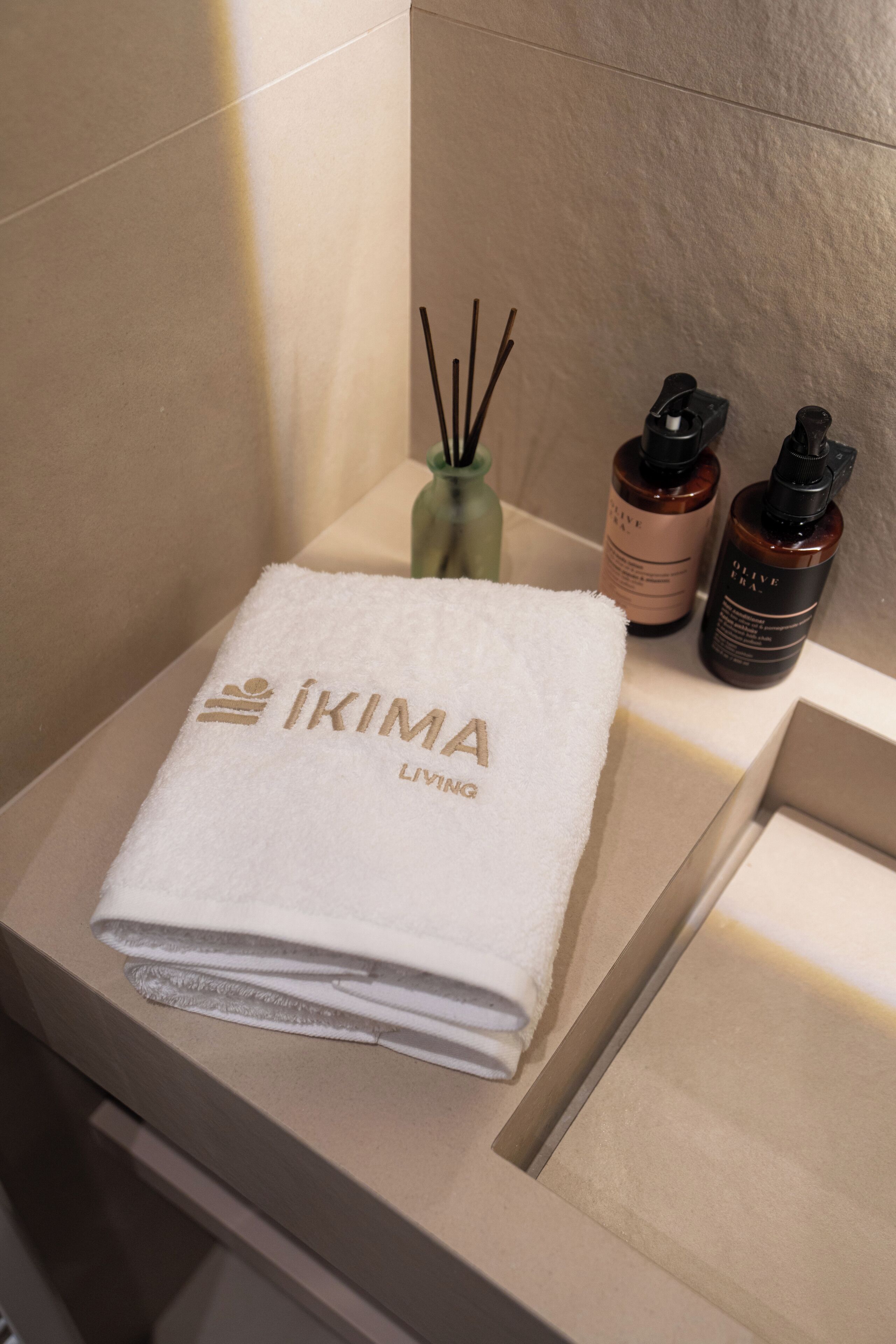 A bathroom with a white towel on a counter, a small vase with reed sticks, and two bottles of olive oil.