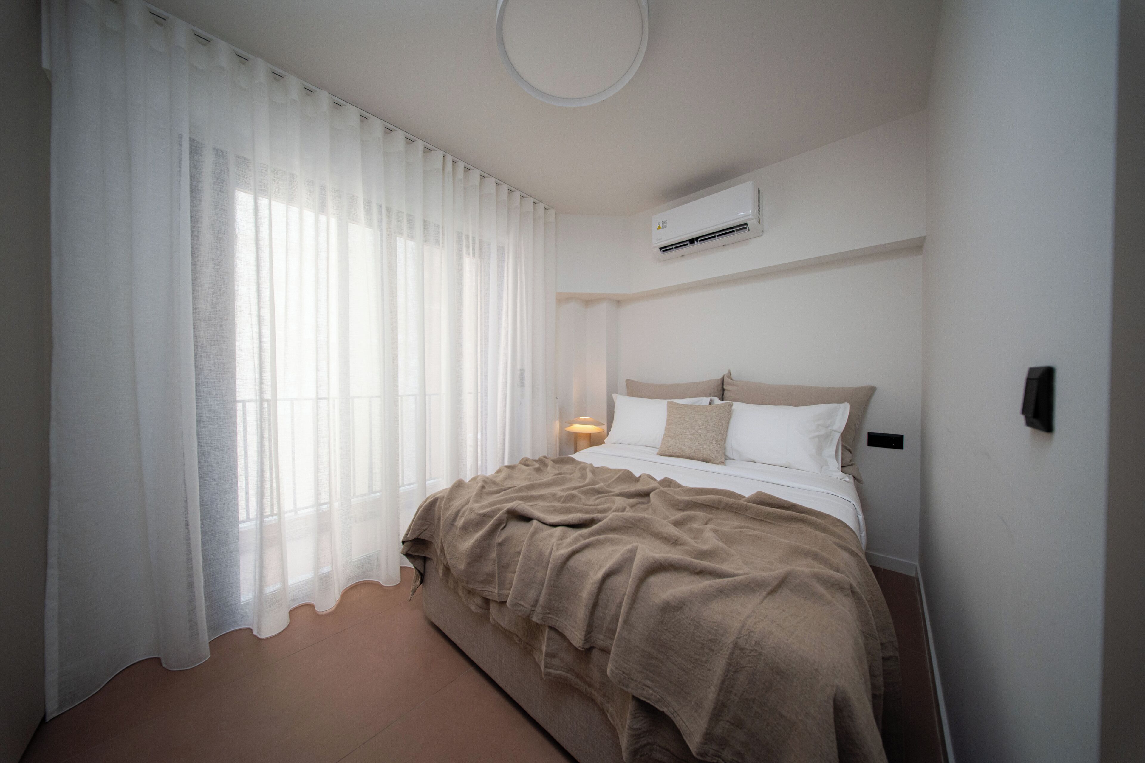 A bedroom with a bed, white curtains, a bedside lamp, and an air conditioning unit.