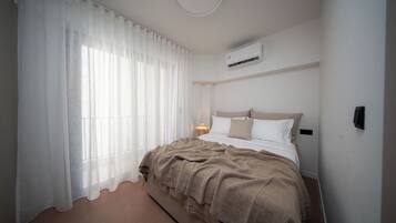 A bedroom with a bed, white curtains, a bedside lamp, and an air conditioning unit.