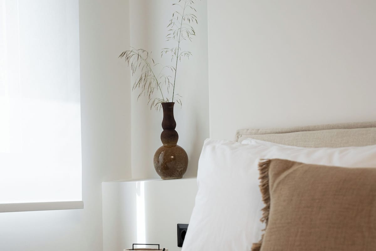 Cama bem arrumada com roupa de cama branca e marrom, um vaso com plantas secas na mesa de cabeceira e um abajur.