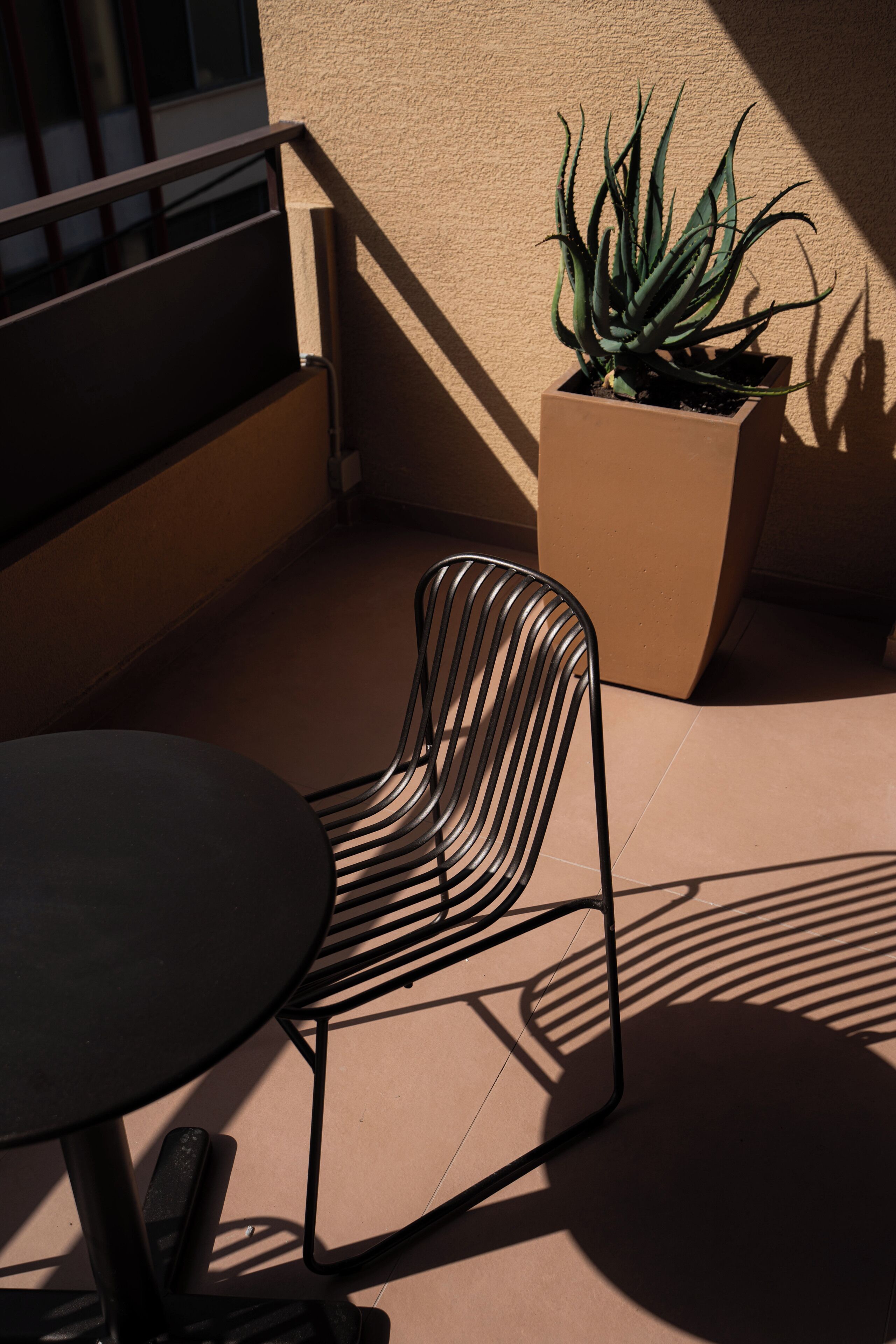 A sunlit outdoor seating area with a metal chair and a table, a potted plant, and a building wall in the background.