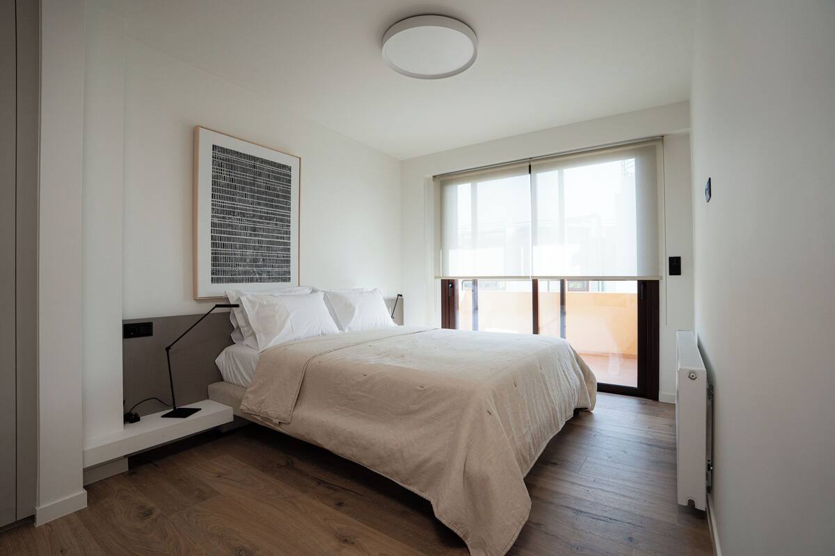 Quarto com cama, mesa de cabeceira, radiador e janela.