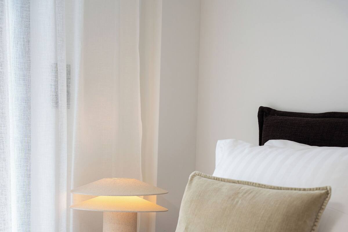 Cama bem arrumada com um cobertor bege, um travesseiro branco e uma cabeceira escura.