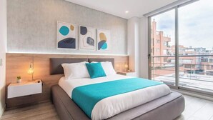 A modern bedroom with a large bed, a nightstand, a bedside table, and a view of buildings through a large window. - Classy 3BR Apartment in Usaquén (Bogotá)