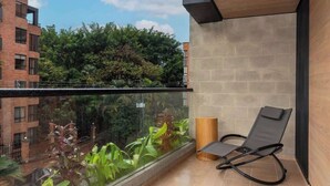 Een balkon met een zwarte lounge stoel, een glazen hekwerk en uitzicht op een bakstenen gebouw en groen.