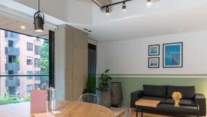 Een moderne woonkamer met een ronde houten tafel, een zwarte bank en een groot raam met uitzicht op een bakstenen gebouw.