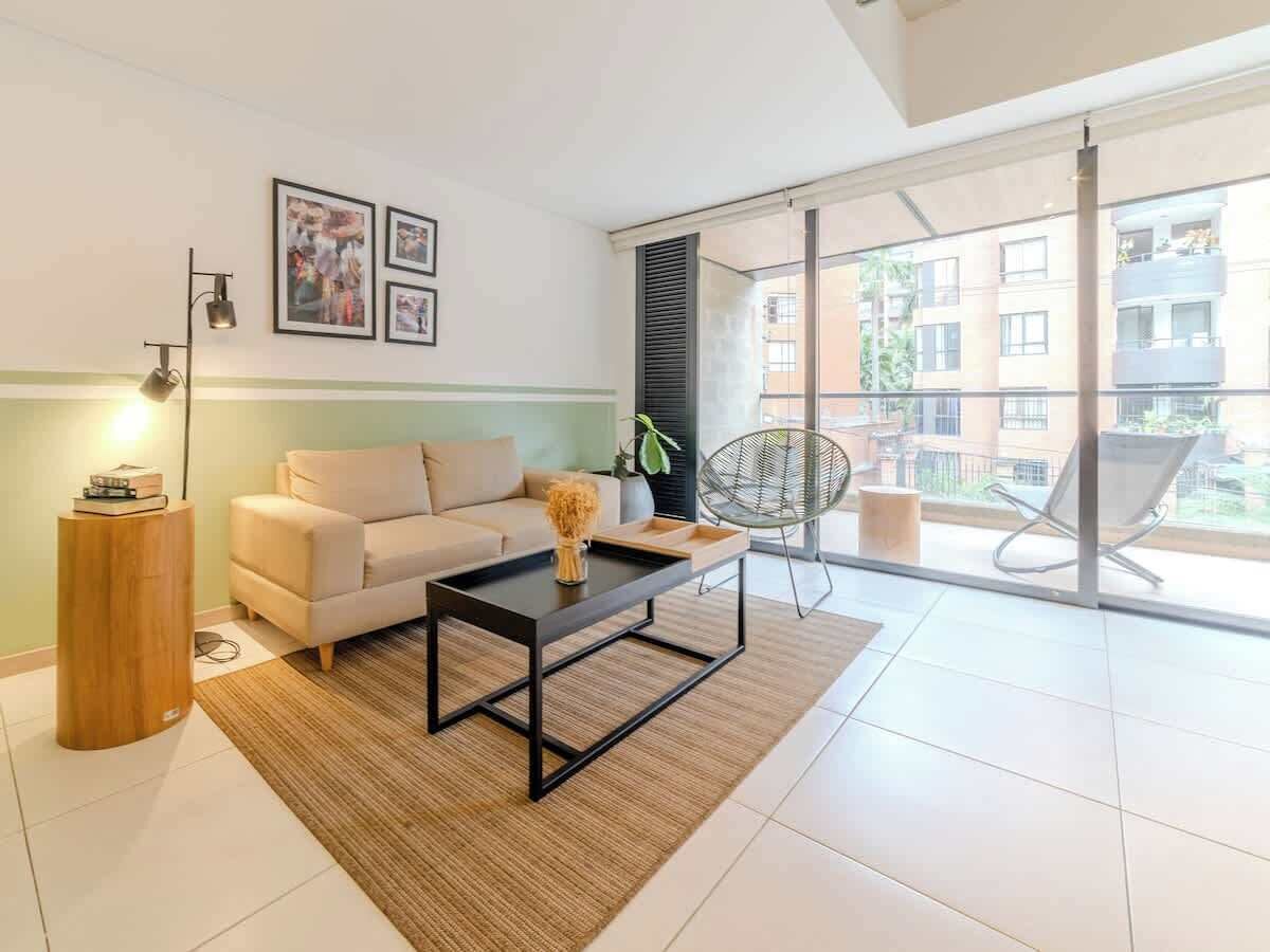 Un salon moderne avec un canapé, une table basse et un fauteuil. Une grande fenêtre offre une vue sur les immeubles.