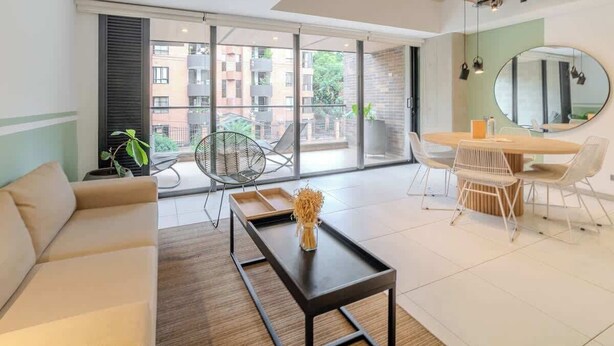 Ein modernes Wohnzimmer mit einer Couch, einem Couchtisch, einem Esstisch mit Stühlen, einem großen Fenster mit Blick auf Gebäude und einem Spiegel an der Wand.