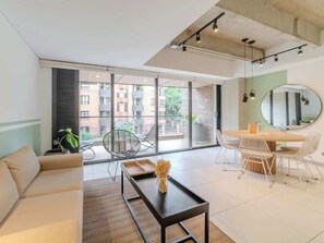 Una sala de estar moderna con sofá, una mesa de centro, una mesa de comedor con sillas, un amplio ventanal con vista a edificios y un espejo en la pared.