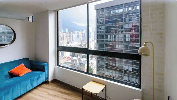 Un salón moderno con un sofá azul, una pequeña mesa de madera y un ventanal con vistas a la ciudad.