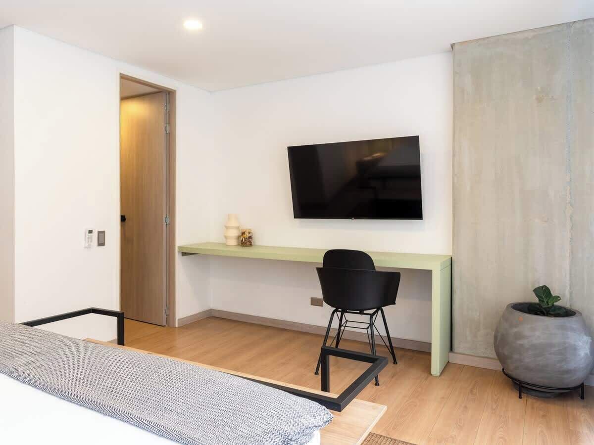 A modern bedroom with a bed, a desk with a chair, a television, and a potted plant.