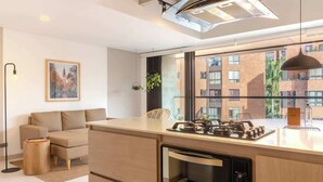 A modern kitchen with a built-in oven, a dining area with a table and chairs, a sofa, and a large window offering a view of a brick building. - Perfect 3 BR w/ Balcony in Laureles Medellín (Medellín)