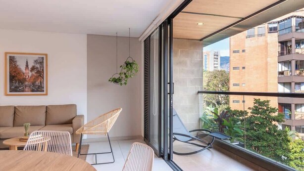 A modern living room with a sofa, chairs, and a dining table. A large window offers a view of the cityscape and greenery. - Perfect 3 BR w/ Balcony in Laureles Medellín (Medellín)