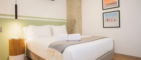 A neatly made bed with white bedding and a grey throw, a bedside table with a lamp, and two framed pictures on the wall.