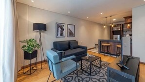 Un salon moderne comprenant un canapé, un fauteuil, une table basse et un téléviseur. Il y a également un coin cuisine avec des tabourets de bar et un réfrigérateur.